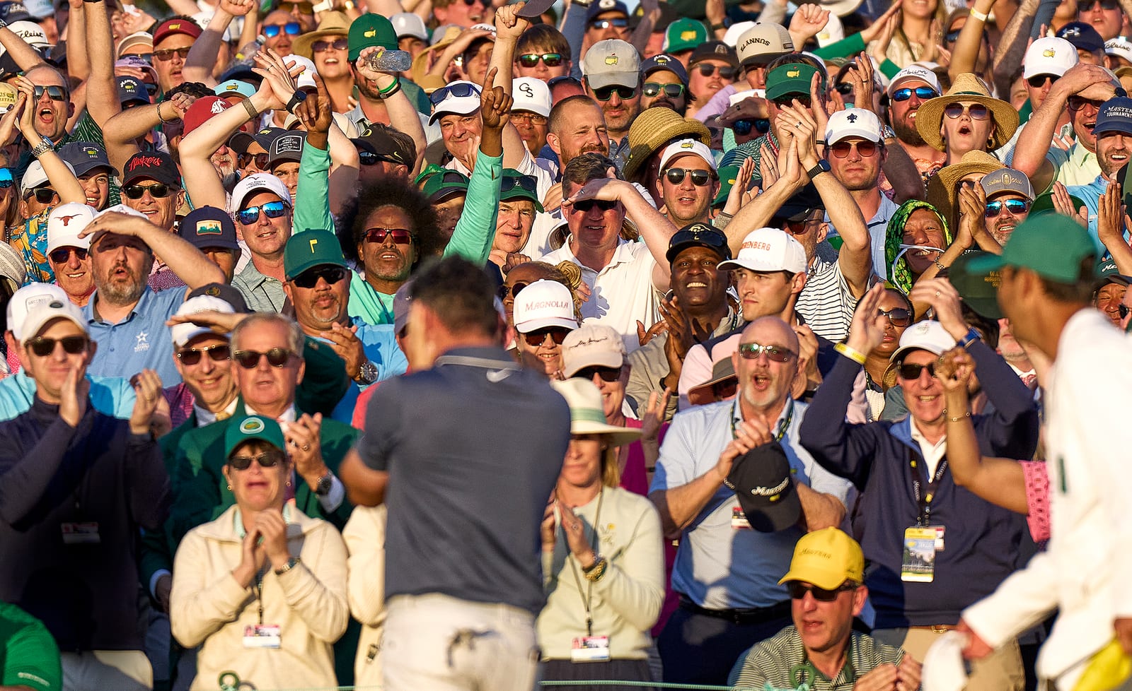 A packed golf gallery cheering as a player walks toward the grandstand in late-afternoon light.
