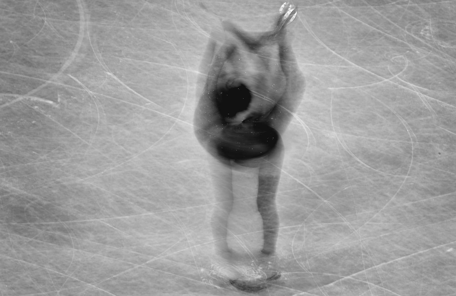 An overhead, motion-blurred black-and-white view of a figure skater spinning on scored ice.